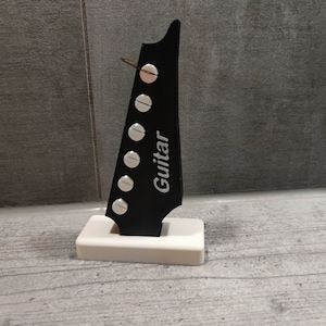 Może przedstawiać: Czarny korpus gitary z srebrnymi kołkami strojeniowymi i napisem "Guitar" nadrukowanym na boku. Korpus jest zamontowany na białej podstawie.