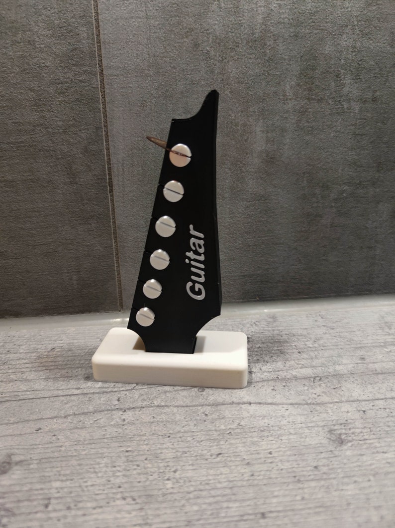 Może przedstawiać: Czarny korpus gitary z srebrnymi kołkami strojeniowymi i napisem "Guitar" nadrukowanym na boku. Korpus jest zamontowany na białej podstawie.