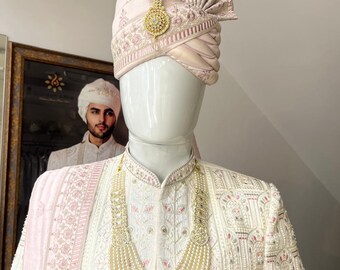 Pode incluir: Um sherwani branco para homem bordado com um dupatta rosa e um colar de pérolas. O sherwani tem um colar alto e mangas compridas. O dupatta é decorado com bordados florais. O colar é feito de pérolas e tem um pingente de ouro.