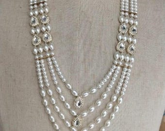 Puede incluir: Un collar de perlas de múltiples hilos con detalles de cristal en forma de lágrima. El collar presenta múltiples hilos de perlas blancas, con grupos de cristales y detalles metálicos dorados. El collar se exhibe en un maniquí.