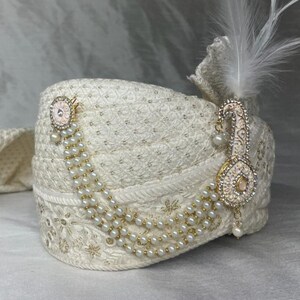 Pode incluir: Um turbante branco com um enfeite dourado e de p&eacute;rolas. O enfeite apresenta um desenho de flor rosa e uma longa corrente de p&eacute;rolas pendentes.