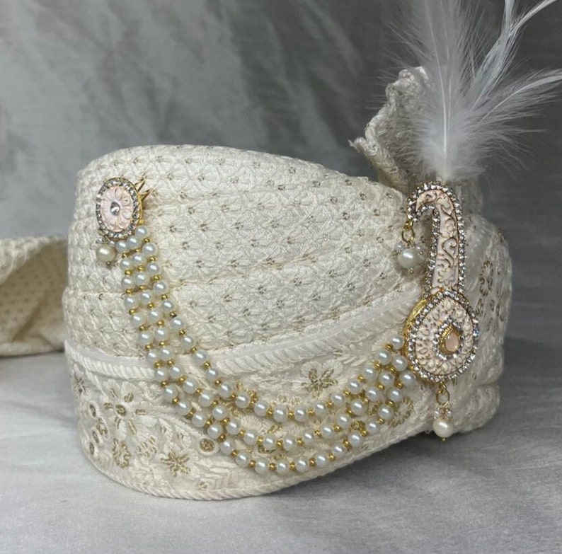 Pode incluir: Um turbante branco com um enfeite dourado e de p&eacute;rolas. O enfeite apresenta um desenho de flor rosa e uma longa corrente de p&eacute;rolas pendentes.