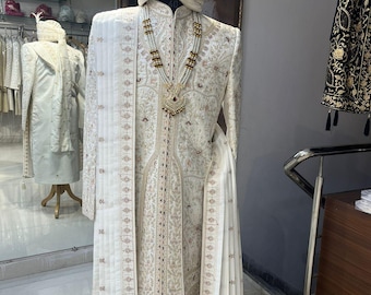 Puede incluir: Un atuendo masculino blanco bordado con una larga chalina blanca, un collar de perlas y un turbante blanco con bordados dorados.