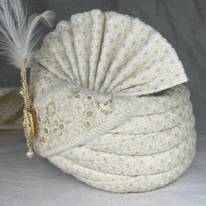 Pode incluir: Um turbante branco com bordados dourados e um adorno de pena branca. O turbante &eacute; feito de um tecido macio e texturizado.