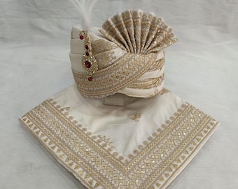 Pode incluir: Um turbante branco com bordados dourados e um detalhe de pena. O turbante é dobrado em forma de leque e tem uma joia vermelha. O turbante é acompanhado por um lenço quadrado branco com bordados dourados.