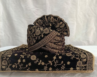 Pode incluir: Um turbante de veludo preto com bordados dourados, com um design floral. O turbante está dobrado e colocado sobre um tecido de veludo preto a condizer com bordados dourados.