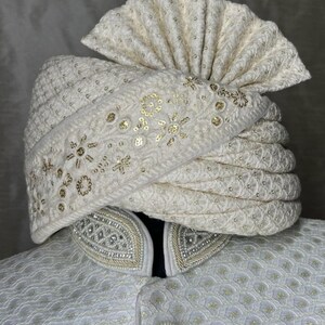 Pode incluir: Um turbante branco com bordados e lantejoulas douradas. O turbante &eacute; dobrado e estilizado com um topo pontudo.