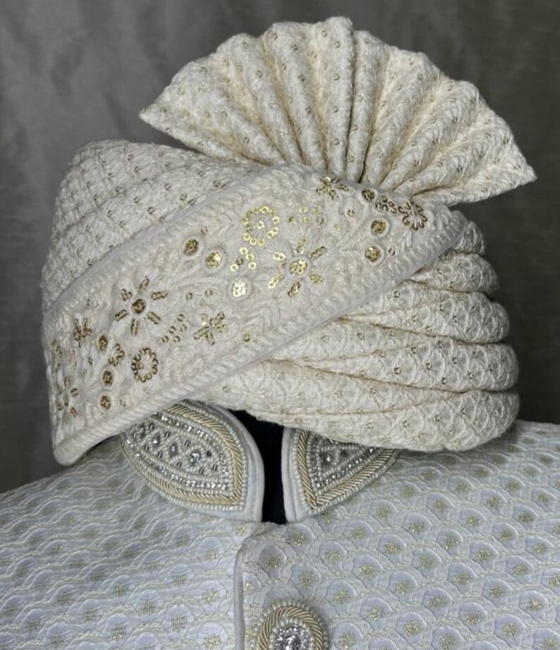 Pode incluir: Um turbante branco com bordados e lantejoulas douradas. O turbante &eacute; dobrado e estilizado com um topo pontudo.