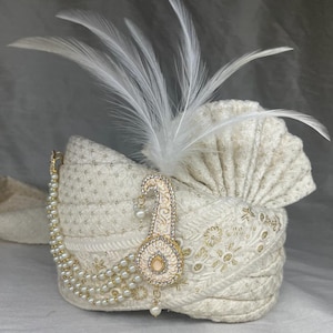 Pode incluir: Um turbante branco com um enfeite de p&eacute;rolas e ouro. O turbante tem uma pena branca no topo.