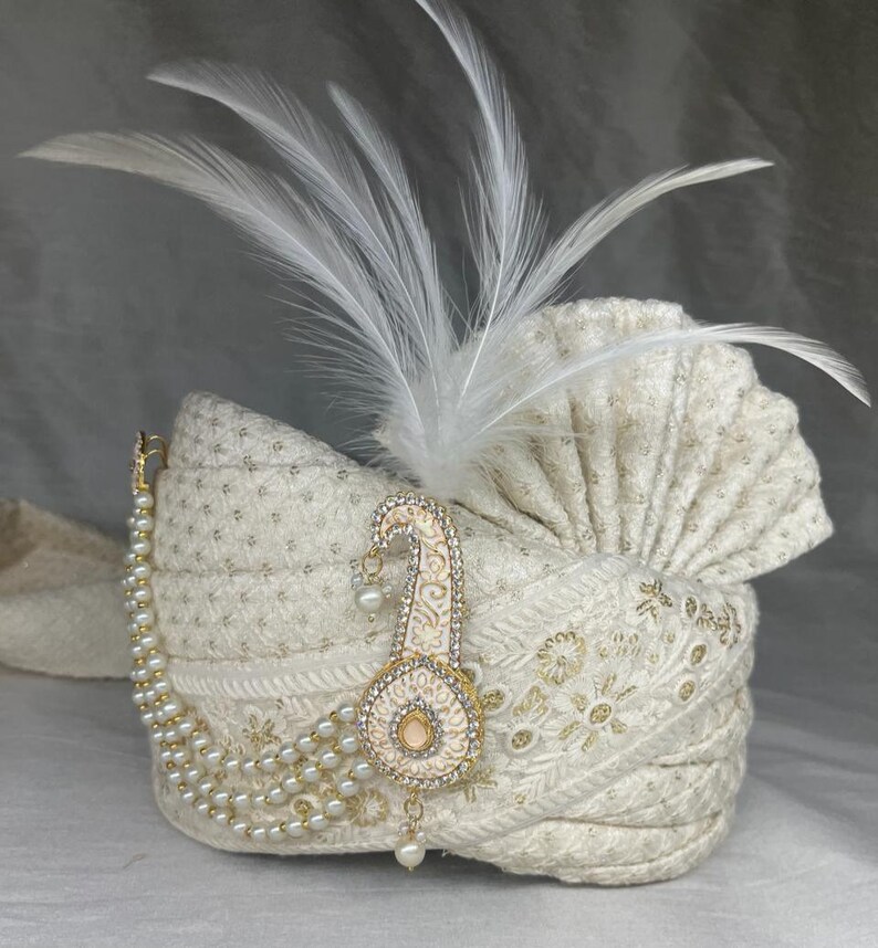 Pode incluir: Um turbante branco com um enfeite de p&eacute;rolas e ouro. O turbante tem uma pena branca no topo.
