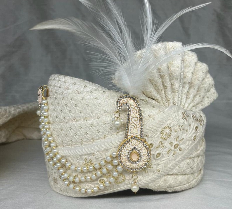 Pode incluir: Um turbante branco com um enfeite de p&eacute;rolas e ouro. O turbante &eacute; decorado com uma pena branca e bordados intrincados.