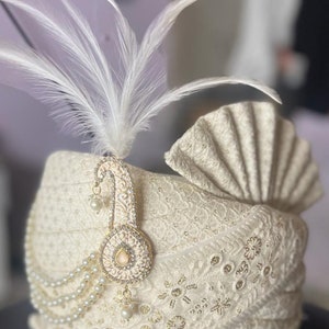 Pode incluir: Um turbante branco com um enfeite de ouro e p&eacute;rolas e uma pena branca.