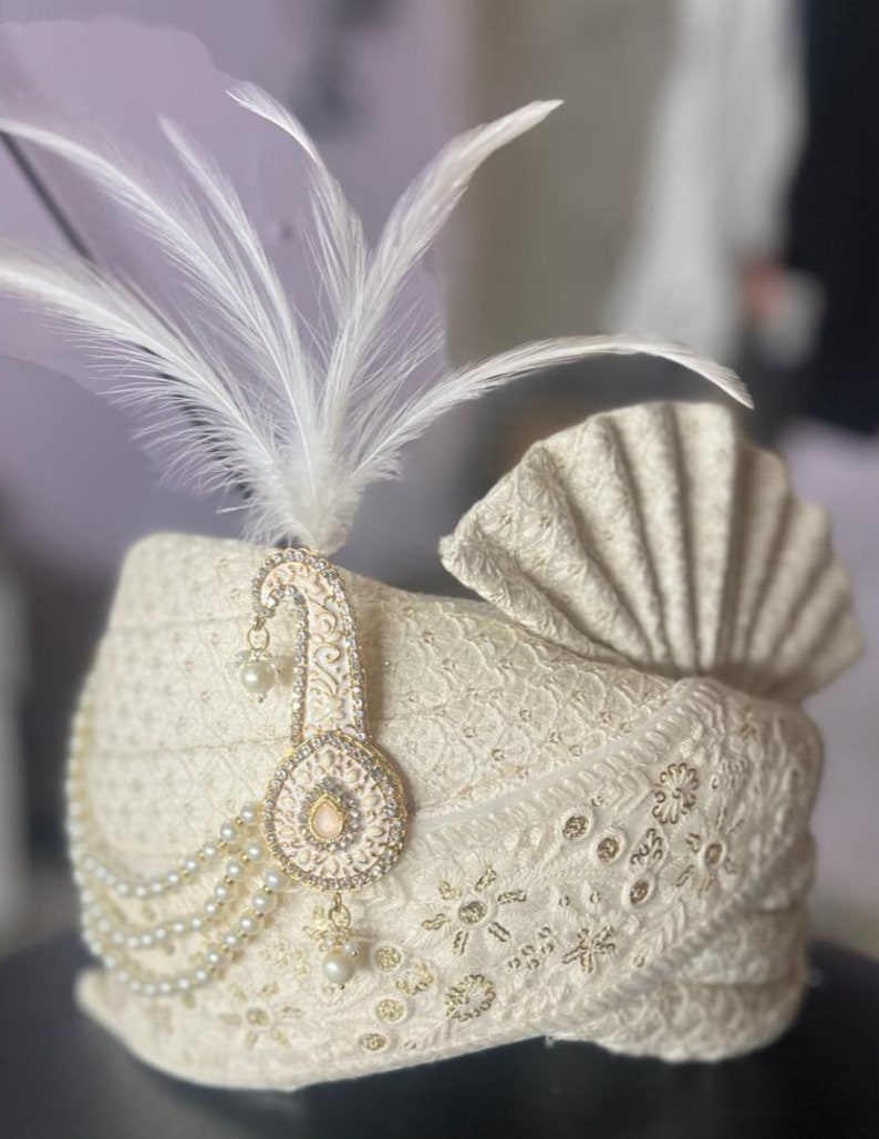 Pode incluir: Um turbante branco com um enfeite de ouro e p&eacute;rolas e uma pena branca.