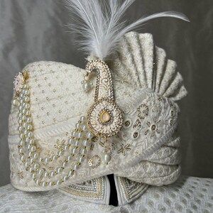 Pode incluir: Um turbante branco com bordados dourados e um colar de p&eacute;rolas. Uma pena branca e um ornamento dourado e branco est&atilde;o presos ao turbante.