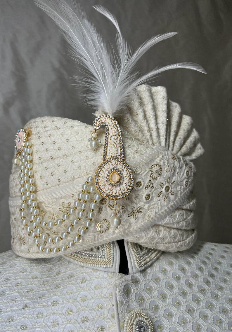 Pode incluir: Um turbante branco com bordados dourados e um colar de p&eacute;rolas. Uma pena branca e um ornamento dourado e branco est&atilde;o presos ao turbante.