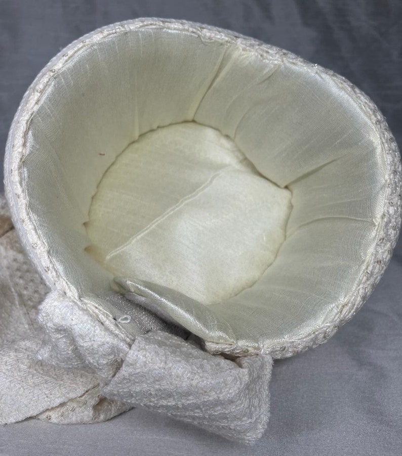 Pode incluir: Um chap&eacute;u branco de croch&ecirc; com forro de cetim branco. O chap&eacute;u tem uma aba larga e um la&ccedil;o de lado.
