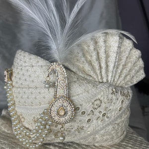 Pode incluir: Um turbante branco com bordados dourados e brancos, uma corrente de p&eacute;rolas e um ornamento branco e dourado com joias e uma pena branca.