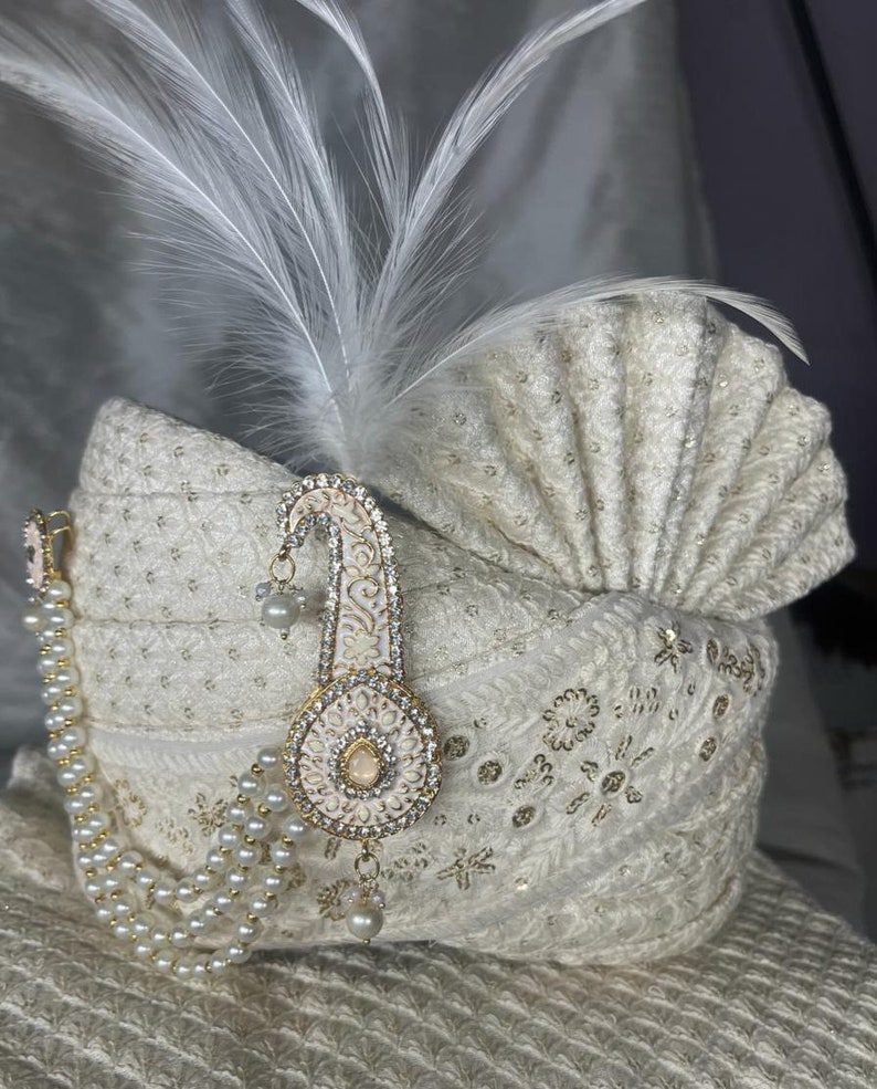 Pode incluir: Um turbante branco com bordados dourados e brancos, uma corrente de p&eacute;rolas e um ornamento branco e dourado com joias e uma pena branca.