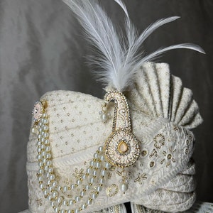 Pode incluir: Um turbante branco com bordados dourados e uma corrente de p&eacute;rolas. Uma pena branca e um ornamento de ouro e rosa com joias est&atilde;o presos ao turbante.