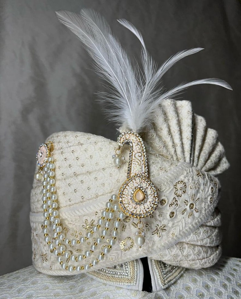 Pode incluir: Um turbante branco com bordados dourados e uma corrente de p&eacute;rolas. Uma pena branca e um ornamento de ouro e rosa com joias est&atilde;o presos ao turbante.