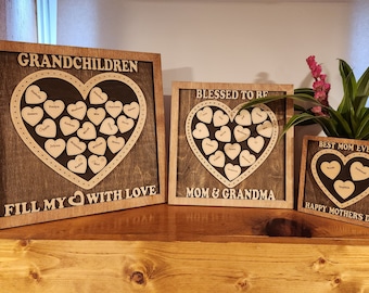 May include: Three wooden framed signs with a heart shape design. The largest sign says "Grandchildren Fill My Heart With Love" and has many small hearts with names inside the large heart. The middle sign says "Blessed To Be Mom & Grandma" and has a smaller heart with names inside. The smallest sign says "Best Mom Ever Happy Mother's Day" and has a heart with three names inside.