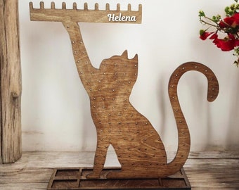 Pode incluir: Suporte de brincos de madeira em forma de gato com uma placa de nome que diz "Helena". O suporte tem uma base com um padrão de grade e uma fileira de pinos para brincos.