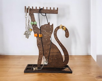 Pode incluir: Um organizador de joias em forma de gato de madeira marrom escuro. O organizador possui uma série de pinos para pendurar colares e uma pequena bandeja na base para guardar anéis e brincos. Várias peças de joias são exibidas.