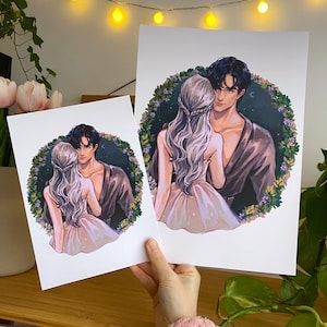 Puede incluir: Dos impresiones artísticas que presentan una ilustración romántica de una pareja abrazándose. La obra de arte representa a un hombre y una mujer dentro de una corona floral, con la mujer con un vestido rosa y el hombre con una bata marrón. Las impresiones son sobre papel blanco.