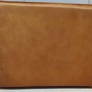 May include: A rectangular, tan leather laptop sleeve. The sleeve has a stitched border and a smooth, slightly textured surface. The leather has a warm, natural color and appears to be well-crafted. The sleeve is designed to protect a laptop.
