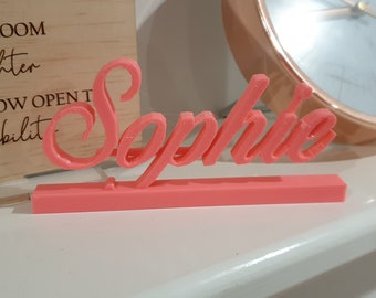 Könnte beinhalten: Ein rosa 3D-gedrucktes Namensschild mit dem Namen "Sophia" in einer kursivschriftlichen Schriftart. Das Schild steht auf einer weißen Oberfläche.