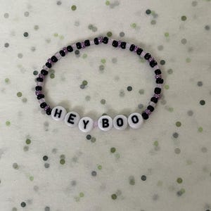Può includere: Un braccialetto di perline con le parole "HEY BOO" scritte con perline di lettere bianche. Il braccialetto presenta perline nere e viola, un accessorio a tema Halloween.