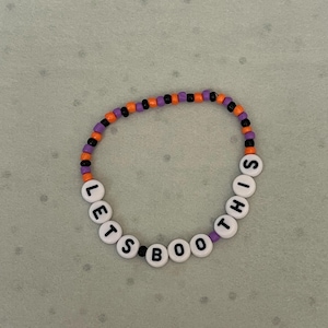 Könnte beinhalten: Ein Perlenarmband mit der Aufschrift "LETS BOO THIS". Das Armband hat ein Halloween-Thema mit orangefarbenen, lila und schwarzen Perlen. Das Armband liegt auf einer hellgrauen Oberfläche.