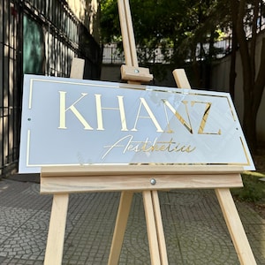 May include: A white sign with gold lettering on a wooden easel. The sign reads "KHANZ Aesthetics".