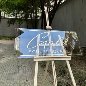 May include: A clear acrylic sign with gold lettering that reads "Gold Sculpt Lounge" on a wooden easel.