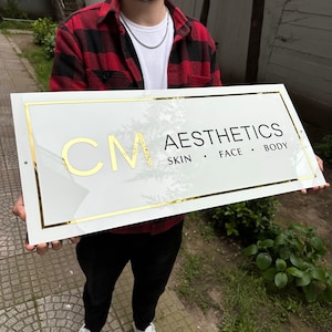 May include: White sign with gold trim and black text. The sign reads "CM AESTHETICS SKIN FACE BODY".