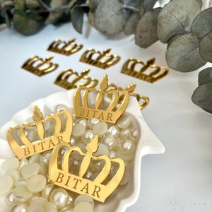 May include: Gold crown-shaped decorations with the name "BITAR" engraved on them. The crowns are scattered on a white surface with white pearl-like beads.