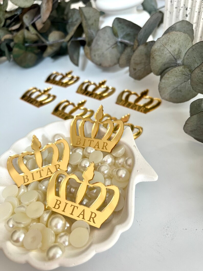 May include: Gold crown-shaped decorations with the name "BITAR" engraved on them. The crowns are scattered on a white surface with white pearl-like beads.