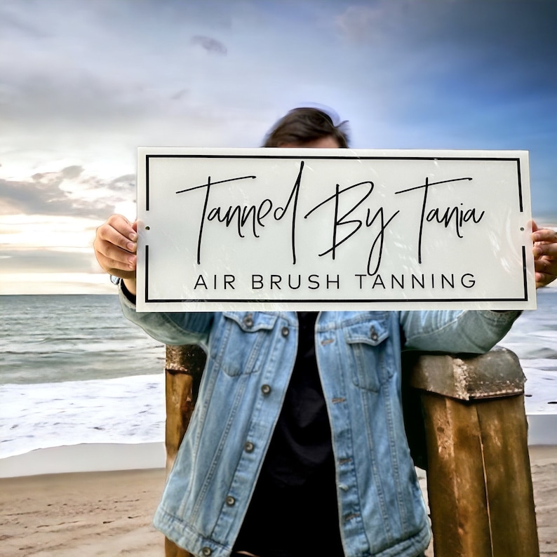 May include: A white sign with black text that reads "Tanned By Tania AIR BRUSH TANNING". The sign is being held by a person in front of a beach scene.