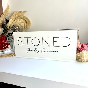 May include: White sign with black text that reads "STONED Jewellery Concierge".