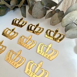 May include: Gold crown-shaped acrylic place cards with the name "BITAR" engraved on each. The crowns are scattered on a white surface.
