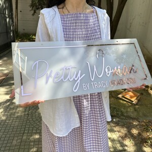 May include: A white sign with a rose gold border and the text "Pretty Woman by Tracy Bradford" in white script.