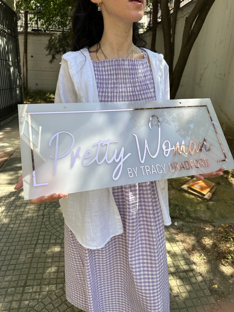 May include: A white sign with a rose gold border and the text "Pretty Woman by Tracy Bradford" in white script.