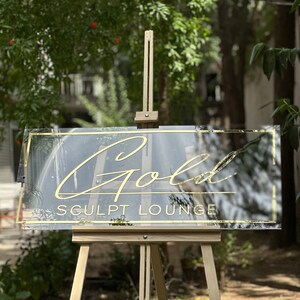 K&ouml;nnte beinhalten: Klarer Acrylschild mit goldfarbenem Schriftzug "Gold Sculpt Lounge". Das Schild hat einen goldenen Rahmen und wird auf einer h&ouml;lzernen Staffelei pr&auml;sentiert. Der Hintergrund zeigt gr&uuml;nes Laub.