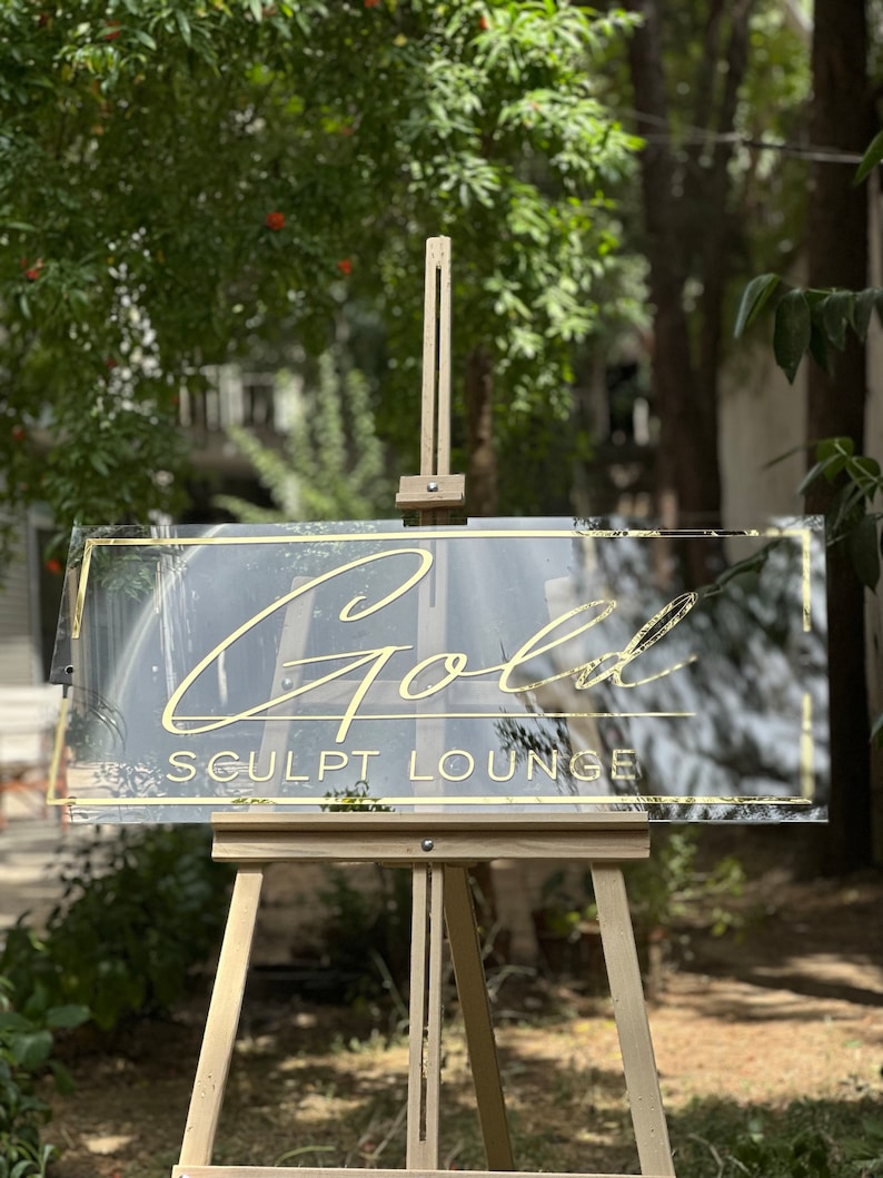 K&ouml;nnte beinhalten: Klarer Acrylschild mit goldfarbenem Schriftzug "Gold Sculpt Lounge". Das Schild hat einen goldenen Rahmen und wird auf einer h&ouml;lzernen Staffelei pr&auml;sentiert. Der Hintergrund zeigt gr&uuml;nes Laub.