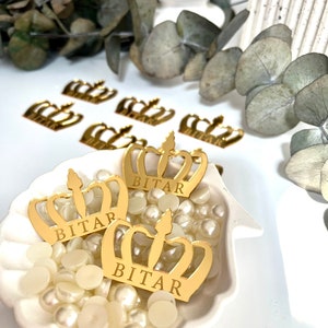 May include: Three gold crown-shaped decorations with the name "BITAR" inscribed on each. The crowns are sitting in a white seashell-shaped dish filled with white pearl-like beads.