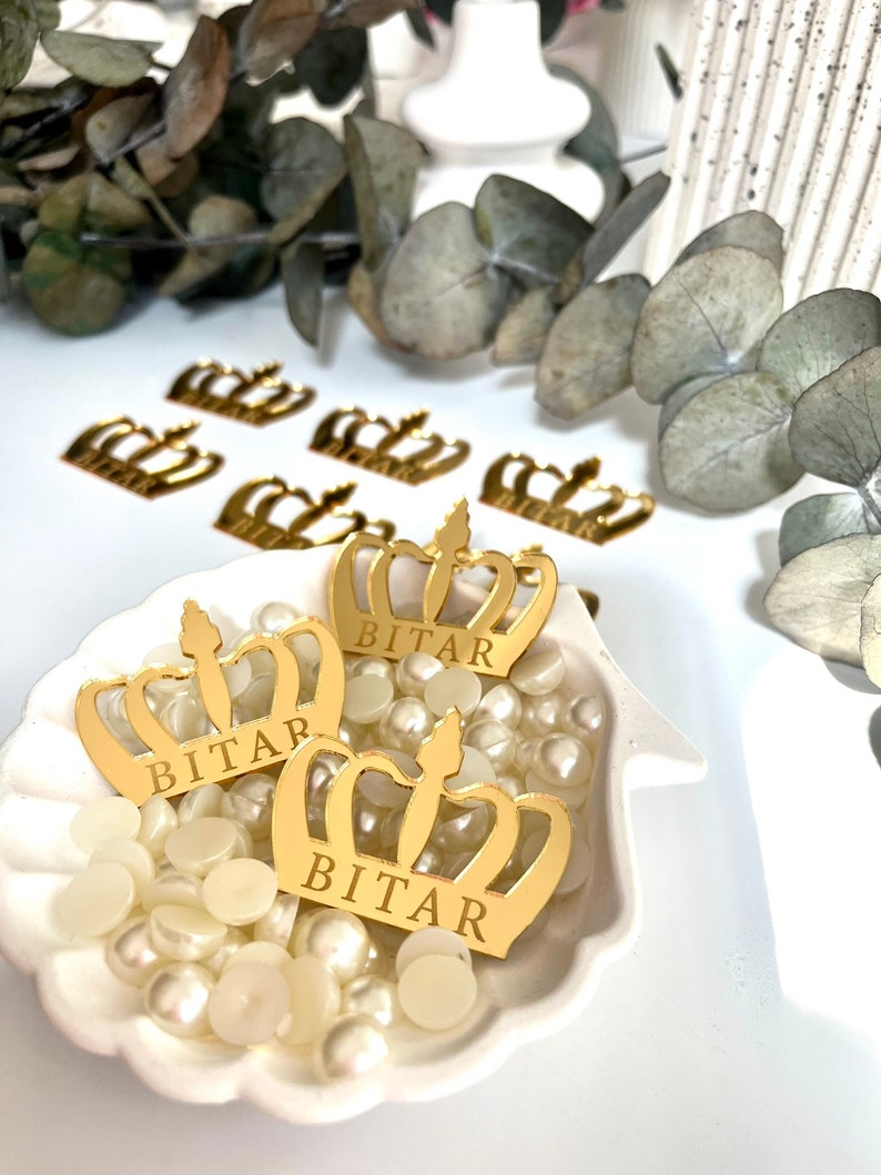 May include: Three gold crown-shaped decorations with the name "BITAR" inscribed on each. The crowns are sitting in a white seashell-shaped dish filled with white pearl-like beads.