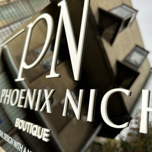 K&ouml;nnte beinhalten: Ein schwarzes Schild mit wei&szlig; erhabener Schrift, das "PN PHOENIX NICHE BOUTIQUE" lautet. Das Schild enth&auml;lt auch den Text "versal design with a niche impact". Das Schild ist an einem Geb&auml;ude angebracht.