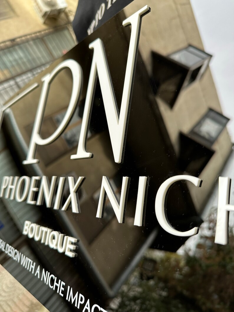 K&ouml;nnte beinhalten: Ein schwarzes Schild mit wei&szlig; erhabener Schrift, das "PN PHOENIX NICHE BOUTIQUE" lautet. Das Schild enth&auml;lt auch den Text "versal design with a niche impact". Das Schild ist an einem Geb&auml;ude angebracht.