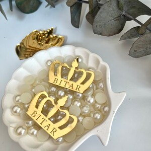 May include: Two gold crown-shaped decorations with the name "BITAR" inscribed on each. The crowns are sitting in a white seashell-shaped dish filled with white pearl-like beads.