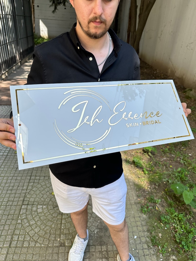 K&ouml;nnte beinhalten: Ein rechteckiges wei&szlig;es Schild mit goldenem Rand und dem Text "Ich Essence SKIN &bull; BRIDAL" in goldfarbenen Buchstaben. Das Schild wird von einer Person gehalten. Das Schild hat ein kreisf&ouml;rmiges Logo-Design.
